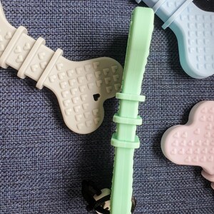 May include: A set of three silicone teethers in the shape of bones. The teethers are in light blue, light green, and light purple. Each teether has a pattern of small hearts on the surface.