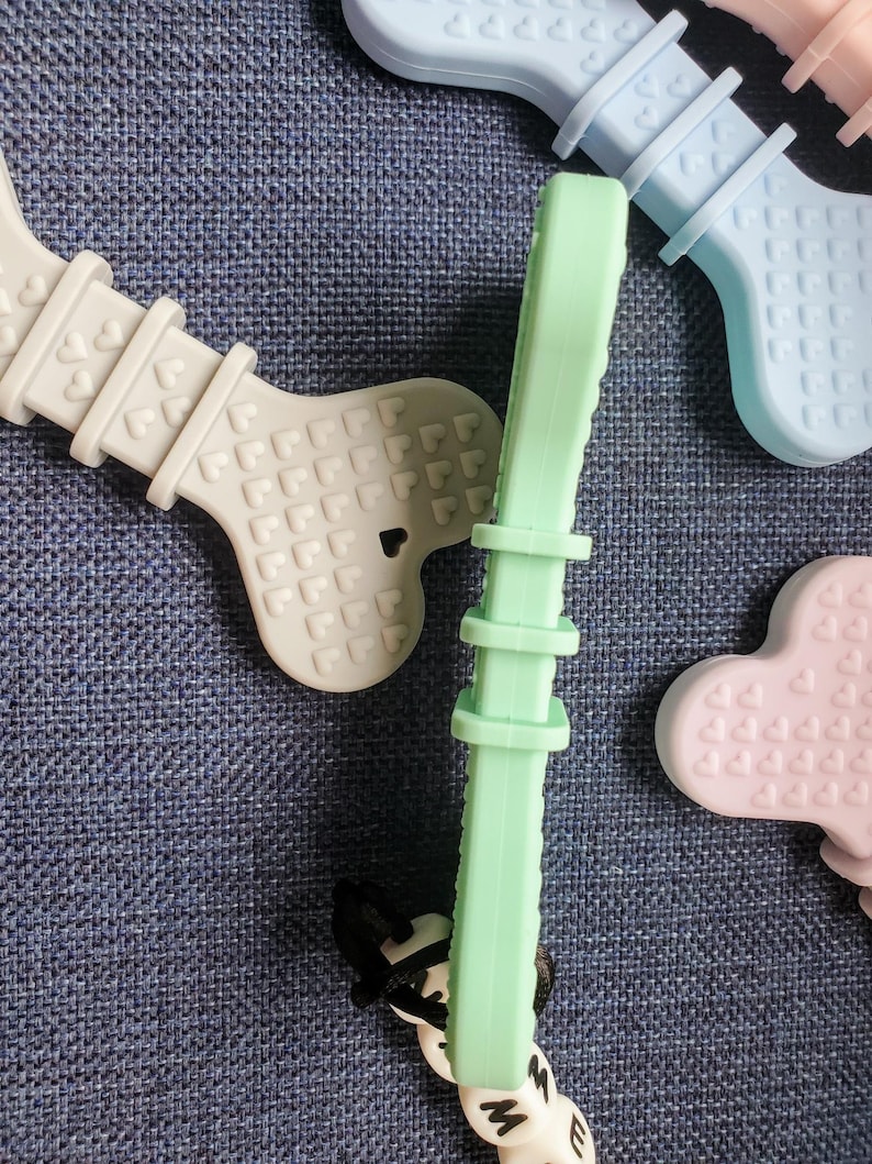 May include: A set of three silicone teethers in the shape of bones. The teethers are in light blue, light green, and light purple. Each teether has a pattern of small hearts on the surface.