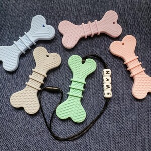 May include: Five silicone teethers shaped like bones in various colors, including light blue, pink, beige, green, and pink. The green teether is attached to a black cord with a white bead necklace that spells "NAME".