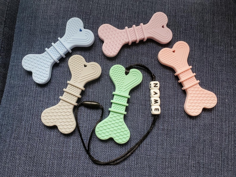 May include: Five silicone teethers shaped like bones in various colors, including light blue, pink, beige, green, and pink. The green teether is attached to a black cord with a white bead necklace that spells "NAME".