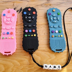May include: Three colorful silicone remote control-shaped baby teethers. One is pink, one is black, and one is blue. Each teether has buttons of various colors and a textured grip. A black cord with the name "NAME" spelled out in white beads is attached.