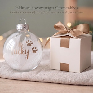 Könnte beinhalten: Eine transparente Glaskugel gefüllt mit weißen Federn, verziert mit dem Schriftzug Lucky und zwei Pfotenabdrücken in Kupfer, inklusive einer quadratischen weißen Geschenkbox mit passendem kupferfarbenem Satinband.