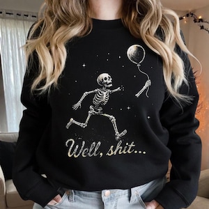 May include: A black long-sleeve sweatshirt featuring a graphic of a skeleton running while holding a balloon. The design includes small white stars and the text "Well, shit..." printed in a cursive font below the skeleton figure.