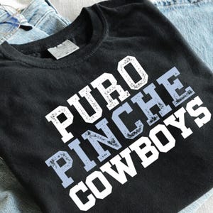 May include: A black short-sleeved t-shirt featuring the text PURO PINCHE COWBOYS printed in a distressed, block-style font. The lettering alternates between white and a light blue gradient, set against the dark fabric of the shirt.