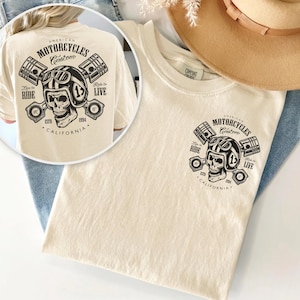 May include: Cream-colored t-shirt featuring a graphic of a skull wearing a motorcycle helmet, pistons, and the text "American Motorcycles Custom." The design includes the words "Ride to Live" and "California 1994."