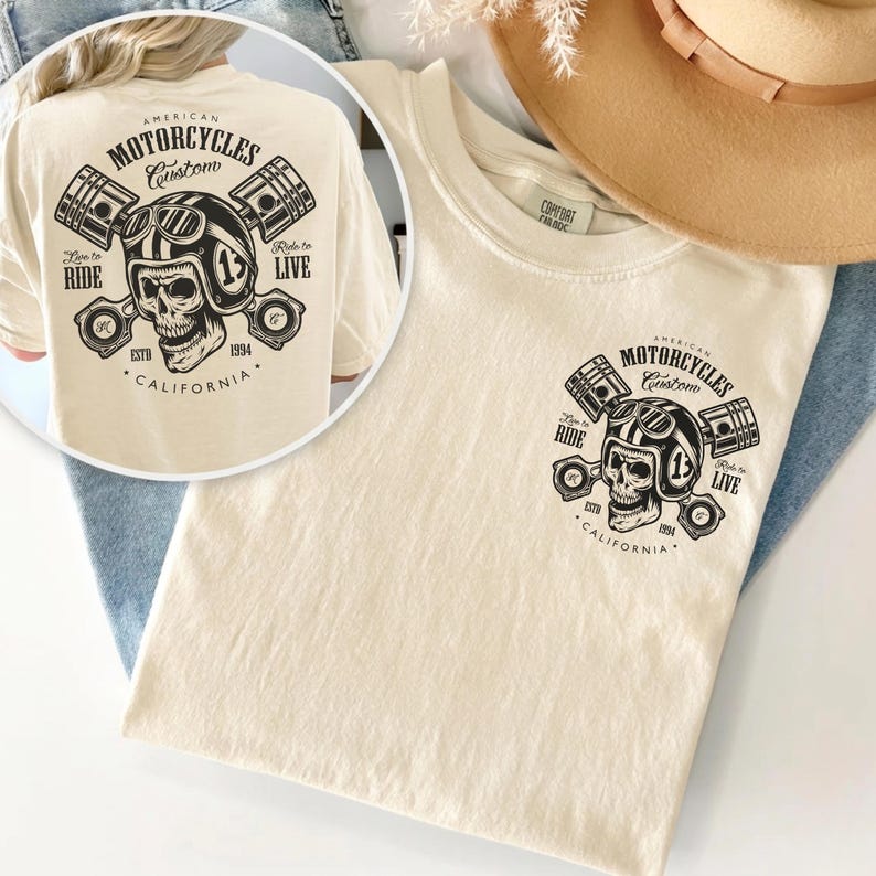 May include: Cream-colored t-shirt featuring a graphic of a skull wearing a motorcycle helmet, pistons, and the text "American Motorcycles Custom." The design includes the words "Ride to Live" and "California 1994."