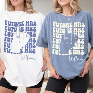 May include: Two short-sleeve t-shirts, one white with blue text and one blue with cream text. Both feature a repeating 'FUTURE MRS' background pattern, a graphic of a hand showing a middle finger with a ring, and the name 'Williams' below.