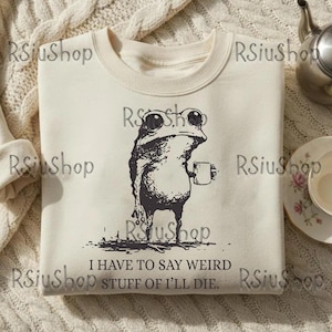 Op de afbeelding: Crèmekleurige sweatshirt met een zwart-wit illustratie van een kikker met een mok. De kikker heeft grote ogen en staat rechtop. De tekst onder de kikker luidt: "I HAVE TO SAY WEIRD STUFF OF I'LL DIE."