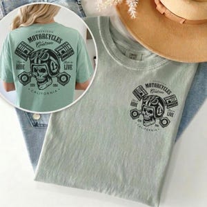 May include: A light green t-shirt featuring a graphic of a skull wearing a motorcycle helmet, pistons, and the text "AMERICAN MOTORCYCLES Custom CALIFORNIA". The shirt has the text "Ride to Live" and "Live to Ride".