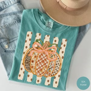 May include: A seafoam green t-shirt featuring a central graphic of a pumpkin decorated with leopard print, floral patterns, and orange gingham. The pumpkin is tied with a peach-colored bow and sits atop vertical stripes with brown polka dots.