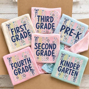 May include: A collection of six folded sweatshirts in cream, pink, and light blue, each featuring a vertical striped pattern with floral, gingham, and star motifs. The text reads FIRST GRADE, THIRD GRADE, PRE-K, SECOND GRADE, FOURTH GRADE, and KINDERGARTEN.