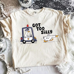 以下が含まれることがあります： クリーム色のTシャツ。漫画のイラストが描かれています。サイレンを鳴らしたパトカーに乗ったガチョウが、逃げる別のガチョウを追いかけています。「GOT TOO SILLY」というテキスト。デザインは黒、白、黄色、赤、青です。
