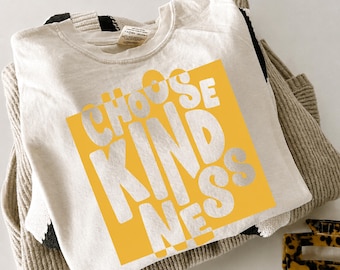 以下が含まれることがあります： クリーム色のTシャツで、大きな黄色の正方形のグラフィックに、太字で波打った歪んだフォントで白い文字「CHOOSE KINDNESS」がプリントされています。Tシャツは、表面に質感のあるベージュと黒のストライプのニットセーターの上に重ねられています。