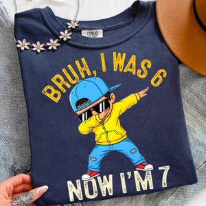 以下が含まれることがあります： ネイビーブルーのTシャツ。サングラス、青いキャップ、黄色のパーカー、青いジーンズを着た、ダブをしている男の子のイラスト。テキストは「Bruh, I was 6 Now I'm 7」と書かれています。