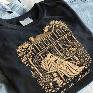 May include: A black t-shirt featuring a light orange graphic print of a two-story house with a ghost sitting in a chair on the porch holding a mug. The design includes trees, flowers, and the text "i am an autumn" written in a cursive script.