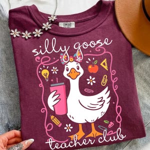 May include: A maroon t-shirt with the words "silly goose teacher club" and a cartoon goose holding a pink tumbler. The goose has a bow and is surrounded by school-themed graphics like an apple, pencil, and lightbulb.