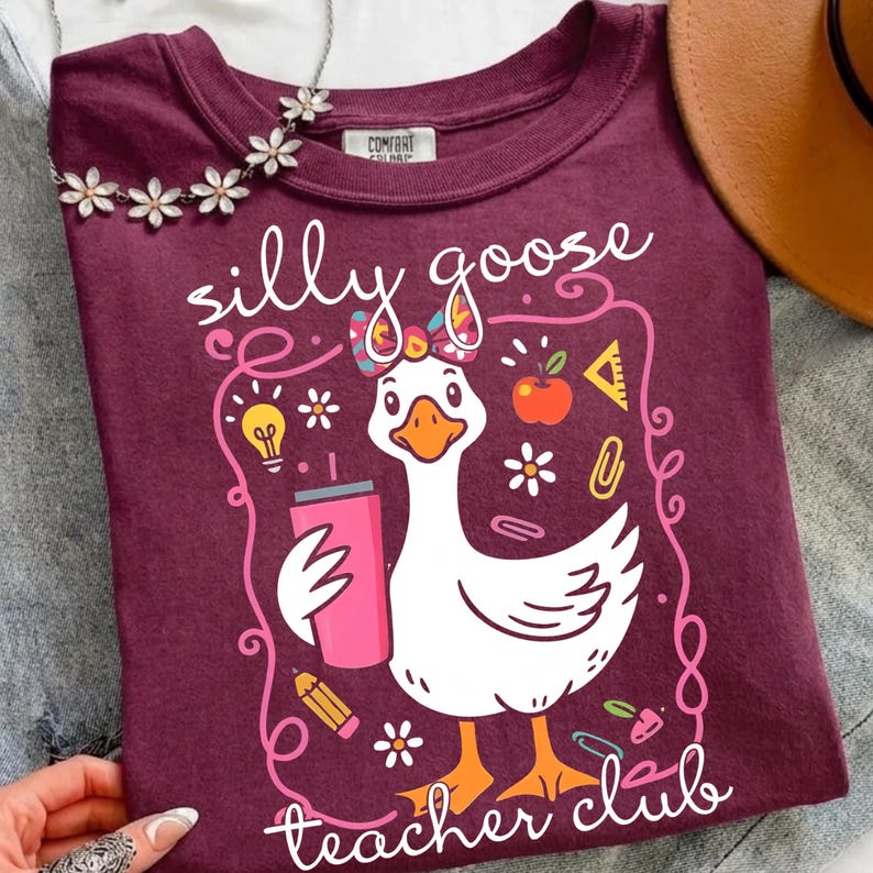 May include: A maroon t-shirt with the words "silly goose teacher club" and a cartoon goose holding a pink tumbler. The goose has a bow and is surrounded by school-themed graphics like an apple, pencil, and lightbulb.
