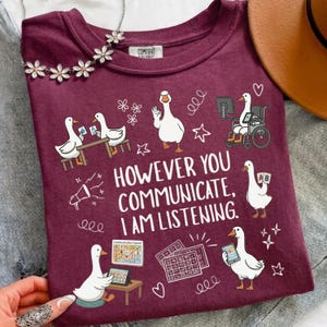 Puede incluir: Una camiseta color granate con ilustraciones blancas de gansos usando varios métodos de comunicación, incluyendo una silla de ruedas, un tablero de comunicación y lenguaje de señas. El texto dice: "HOWEVER YOU COMMUNICATE, I AM LISTENING."