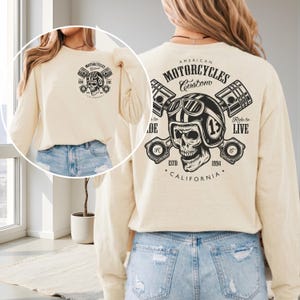 May include: Cream-colored long-sleeve shirt with a motorcycle-themed graphic. The design features a skull wearing a helmet, pistons, and the text "American Motorcycles Custom California". The shirt has a crew neck and a relaxed fit.