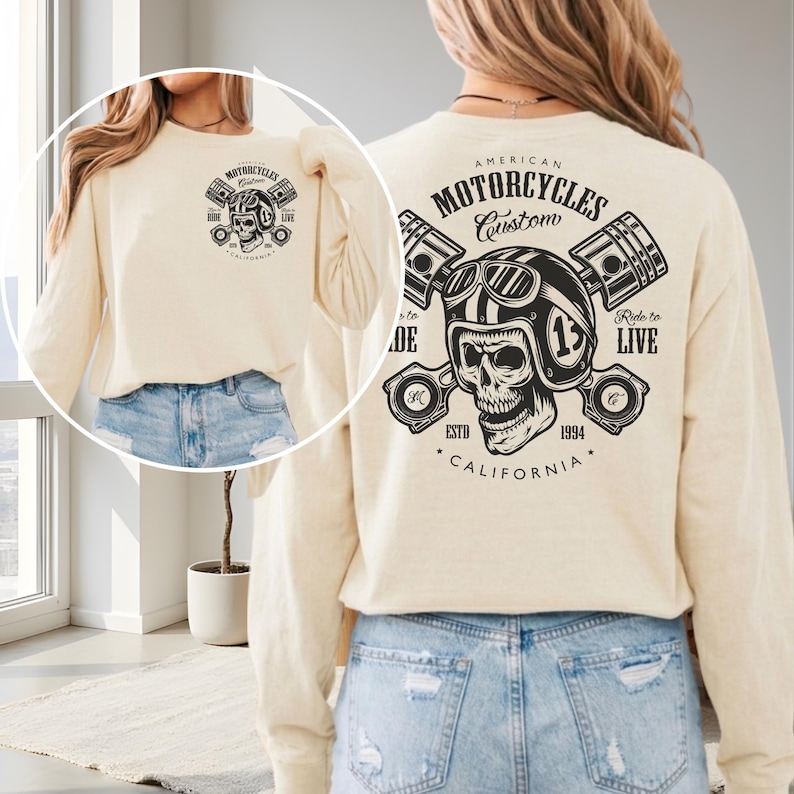 May include: Cream-colored long-sleeve shirt with a motorcycle-themed graphic. The design features a skull wearing a helmet, pistons, and the text "American Motorcycles Custom California". The shirt has a crew neck and a relaxed fit.