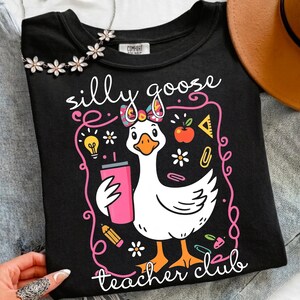 May include: Black t-shirt with a whimsical design featuring a white goose holding a pink tumbler. The text "silly goose teacher club" is displayed in a playful font. The design includes school-themed elements like an apple, pencil, and lightbulb.