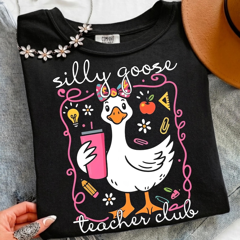May include: Black t-shirt with a whimsical design featuring a white goose holding a pink tumbler. The text "silly goose teacher club" is displayed in a playful font. The design includes school-themed elements like an apple, pencil, and lightbulb.
