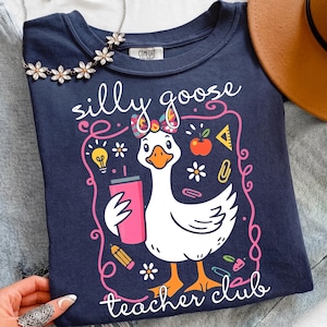 May include: Navy blue t-shirt with a whimsical design featuring a cartoon goose holding a pink tumbler. The text reads "silly goose teacher club" in a playful font, surrounded by school-themed illustrations.