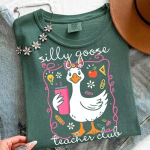 May include: A dark green t-shirt with a whimsical graphic of a goose wearing a bow and holding a pink tumbler. The text "silly goose teacher club" is displayed in a playful font, surrounded by school-themed illustrations.