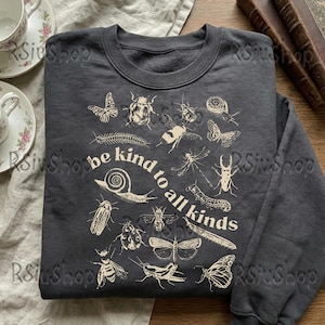 May include: Charcoal grey sweatshirt featuring a cream insect illustration and the text "be kind to all kinds." The design includes butterflies, beetles, and bees. The crew neck sweatshirt is made of a soft material.