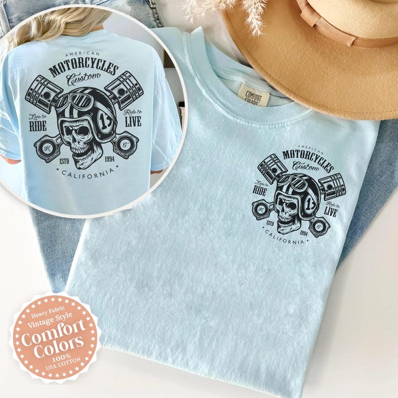 May include: Light blue t-shirt with a graphic featuring a skull wearing a motorcycle helmet, pistons, and the text "AMERICAN MOTORCYCLES Custom CALIFORNIA." The shirt also has the text "Ride to Live" and "How to Ride."