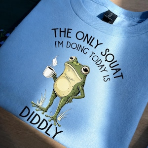 Könnte beinhalten: Ein hellblaues Sweatshirt mit Rundhalsausschnitt und einer Grafik eines grünen Frosches, der eine dampfende weiße Kaffeetasse hält. Der schwarze Text über und unter dem Frosch lautet, THE ONLY SQUAT I'M DOING TODAY IS DIDDLY. Gerippter Kragen.