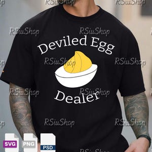 Può includere: T-shirt nero con la scritta bianca "Deviled Egg Dealer" e una grafica di uovo alla diavola. La grafica dell'uovo presenta un tuorlo giallo e un albume bianco. La maglietta è in stile casual.