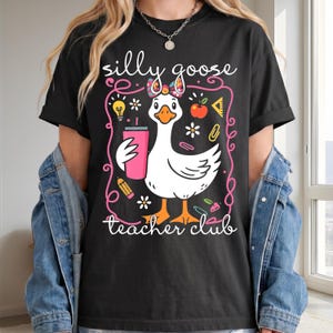 May include: Black t-shirt with a whimsical graphic of a white goose holding a pink tumbler, surrounded by school-themed illustrations. The text "silly goose teacher club" is written in white script.