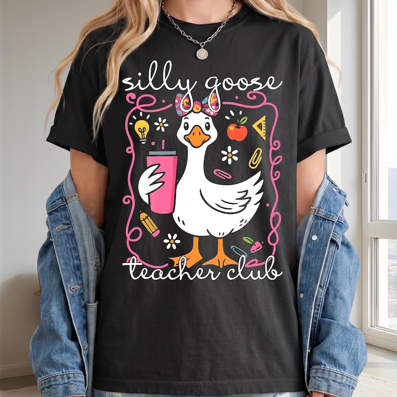 May include: Black t-shirt with a whimsical graphic of a white goose holding a pink tumbler, surrounded by school-themed illustrations. The text "silly goose teacher club" is written in white script.