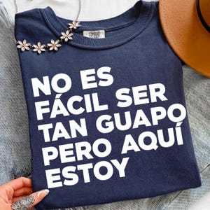 May include: A navy blue t-shirt featuring the white printed text NO ES FÁCIL SER TAN GUAPO PERO AQUÍ ESTOY. The shirt has a crew neckline with a Comfort Colors brand label inside and is decorated with a silver-toned floral chain necklace.