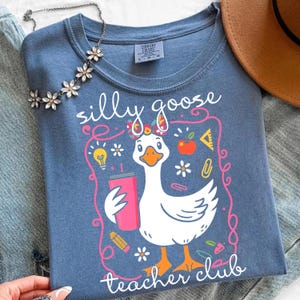 May include: A blue Comfort Colors t-shirt featuring a cartoon goose holding a pink tumbler. The shirt has the words "silly goose teacher club" in white script. The design includes school-themed graphics like an apple, pencil, and lightbulb.