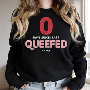 May include: A black long-sleeve crewneck sweatshirt featuring a large red number zero at the top, followed by the white text "DAYS SINCE I LAST" and the pink text "QUEEFED" with a small white zigzag line underneath the word.