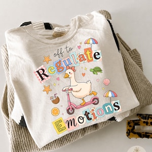Pode incluir: Uma t-shirt cor de creme com um gráfico de um ganso a andar de trotinete cor-de-rosa. O texto "off to Regulate Emotions" está escrito em letras de bloco coloridas e estampadas. O design inclui uma tartaruga, estrela-do-mar, concha, bebida de coco, fatia de laranja e dois guarda-sóis coloridos.