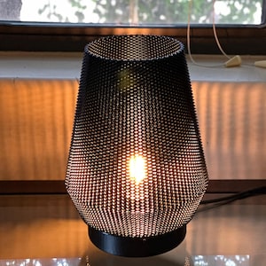 May include: A black metal table lamp with a mesh shade. The lamp is turned on and the light shines through the mesh.