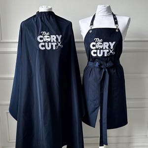 Puede incluir: Una capa de barbero y un delantal azul marino con el texto "The Cory Cut" y un pequeño gráfico de autocaravana. La capa tiene un ribete azul y el delantal tiene una corbata negra.
