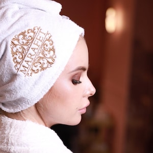 Może przedstawiać: Biały szlafrok kąpielowy z białym ręcznikiem owiniętym wokół głowy. Ręcznik ma haftowany złoty napis "MARAFEAT".