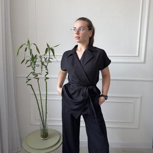 Pode incluir: Um top preto envolvente com mangas curtas e um par de calças de perna larga a condizer. O top tem um fecho de laço na cintura. A roupa é usada com um relógio. Uma pequena mesa lateral com uma planta está no fundo.