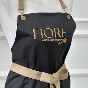 May include: Black apron with gold embroidered text "FIORE". It features a beige waist tie and adjustable straps with gold-coloured metal hardware. The text "ключ до серця" and a key graphic are also embroidered in gold.