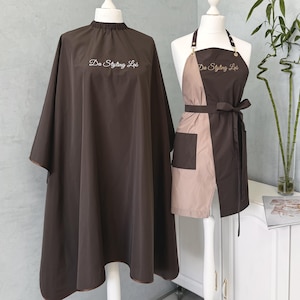 Op de afbeelding: Twee bruine salon capes en schorten op mannequins. De cape is effen bruin met de tekst "Da Styling Lab" geborduurd op de voorkant. Het schort is tweekleurig, bruin en beige, met een zak en dezelfde geborduurde tekst.