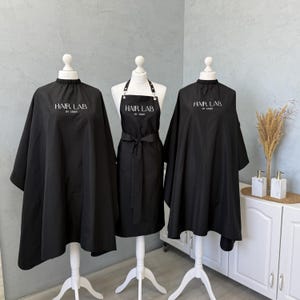 Puede incluir: Dos capas de peluquería negras y un delantal negro sobre maniquíes blancos. Cada artículo presenta el texto blanco "HAIR LAB BY JENNY" impreso en el área del pecho. Las capas tienen un cuello fruncido, mientras que el delantal tiene tiras ajustables.