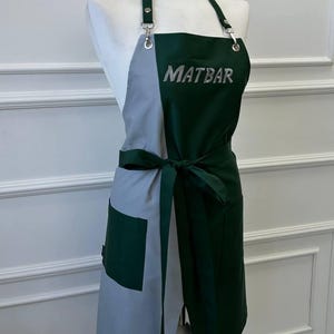 May include: A two-tone apron with a dark green and gray design. The apron features the word "MATBAR" in silver lettering. It has a pocket on the gray side and a tie-waist design. The apron is displayed on a mannequin.
