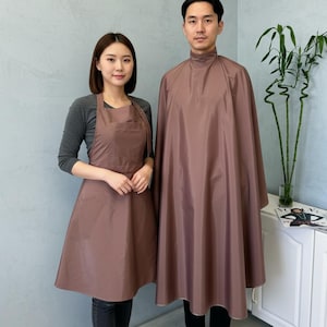 Può includere: Due mantelli e grembiuli da salone marroni. Il grembiule ha un bavaglino e un cinturino in vita. La mantella è un design a copertura totale con un collo alto. Entrambi sono realizzati in un tessuto liscio e tinta unita. Lo sfondo è un muro grigio chiaro.
