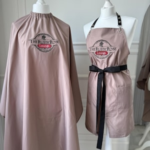 Può includere: Due mantelle e grembiuli da salone color talpa su manichini. La mantella è un design a copertura totale, mentre il grembiule ha un fiocco nero e una tasca. Entrambi gli articoli presentano il testo "THE RUSTIC ROSE Lounge" in un logo circolare.