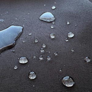 May include: Close-up of a dark gray fabric surface with multiple water droplets, some of which are large and round, and others are smaller and more dispersed.