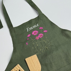 Puede incluir: Un delantal de color verde oliva con un diseño estampado de flores rosas con tallos y hojas verdes. El nombre Emma está impreso en letras blancas sobre el arreglo floral. Dos utensilios de cocina de madera clara son visibles.